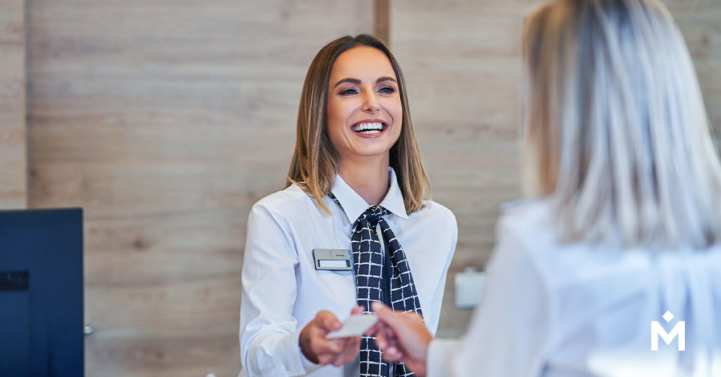 A woman hospitality worker stands behind the front desk of a hotel, smiling and handing back a credit card to a guest